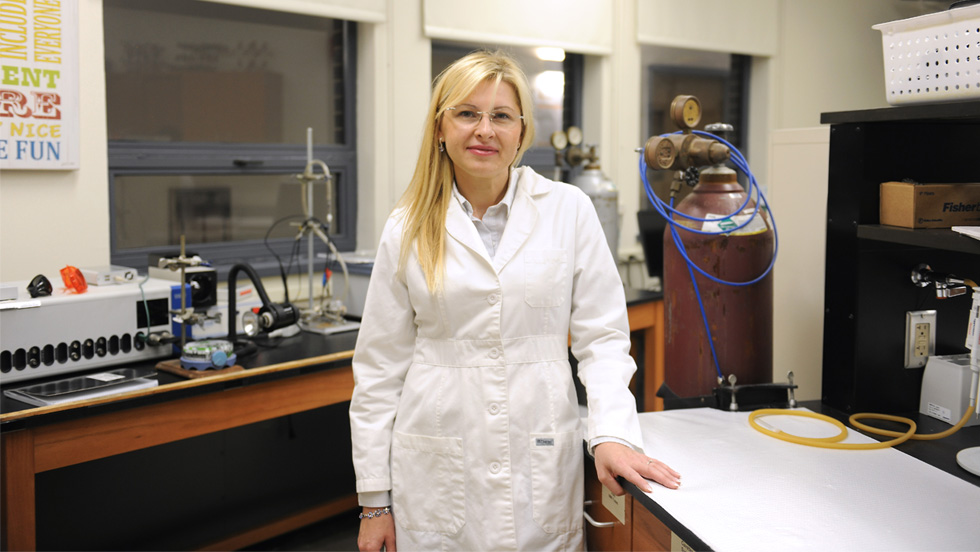 A woman with blond hair and glasses, wearing a white lab coat, in a chemistry lab