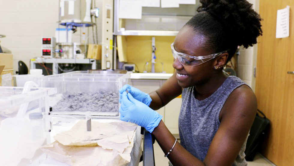Keanna Jardine wears protective glasses and gloves as she handles a gray substance similar to moon rocks.