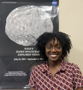 Dr. Jardine stands in front of a poster that says "NASA's Dawn Spacecraft Explores Vesta: July 16, 2011– September 5, 2012." The poster includes a large picture of the asteroid Vesta, which is the second most massive object in the asteroid belt.