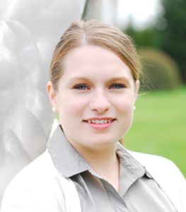 A woman grinning with dirty blonde that's hair tied back. She wears a gray button up with a white sweater. She stands in front of a blurred background that is half gray and half green.