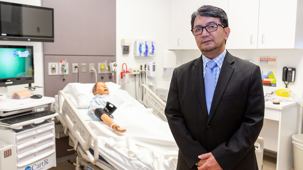 Professor Pajarillo stands in the College of Nursing and Public Health's simulation lab, a hospital-like setting for nurse training. Behind him is a manikin in a hospital bed.