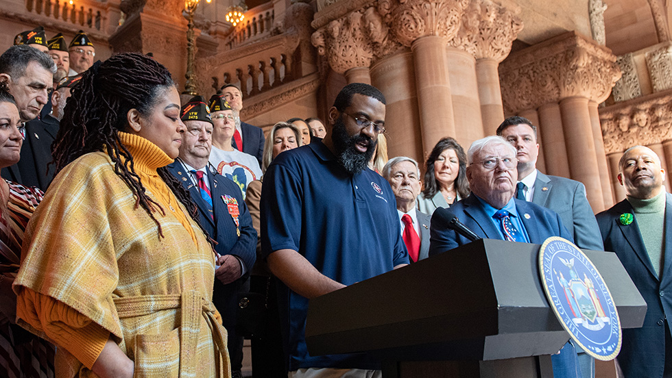 Walters stands at the microphone on a pedestal fronted by the seal of the State of New York. He is surrounded by veterans, many wearing American Legion caps. The scene is inside the ornate lobby of the historic state capitol.  