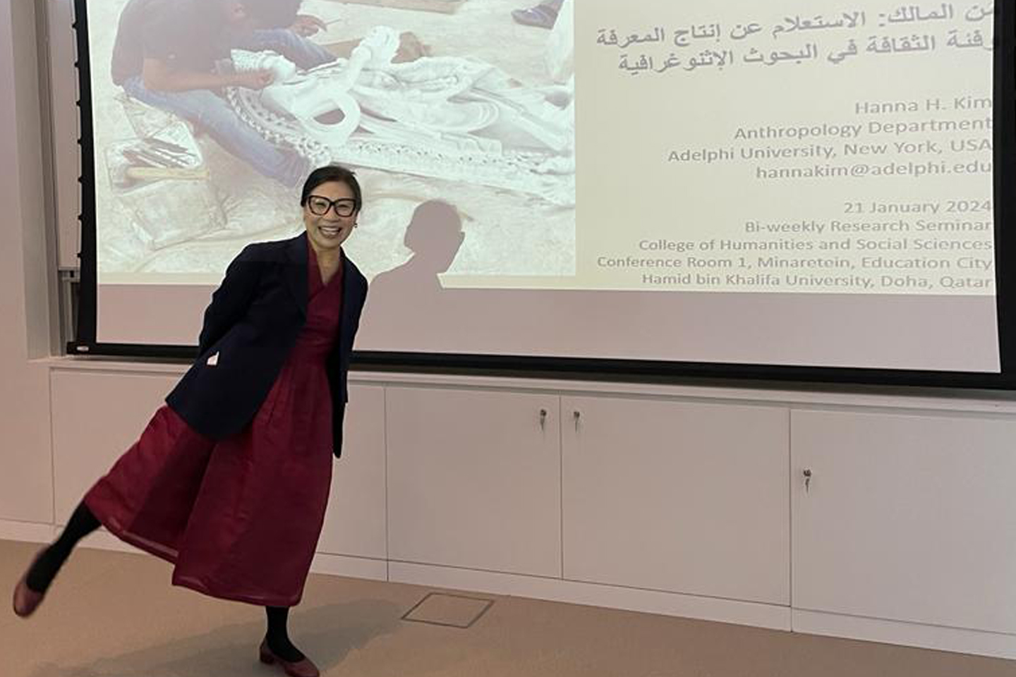 Professor Hanna Kim is in red dress and stands with one leg out to the side. Her arms are behind her back. She has large led framed glasses and is grinning.