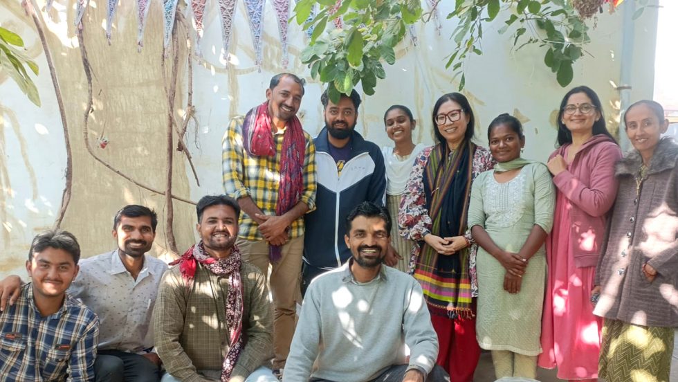 Professor Hanna Kim, PhD (fourth from the right, wearing eyeglasses and traditional Hindu garb), stands under a tree and in front of a mural of a tree, along with six men and four women, some of whom also are wearing Hindu apparel or scarves.