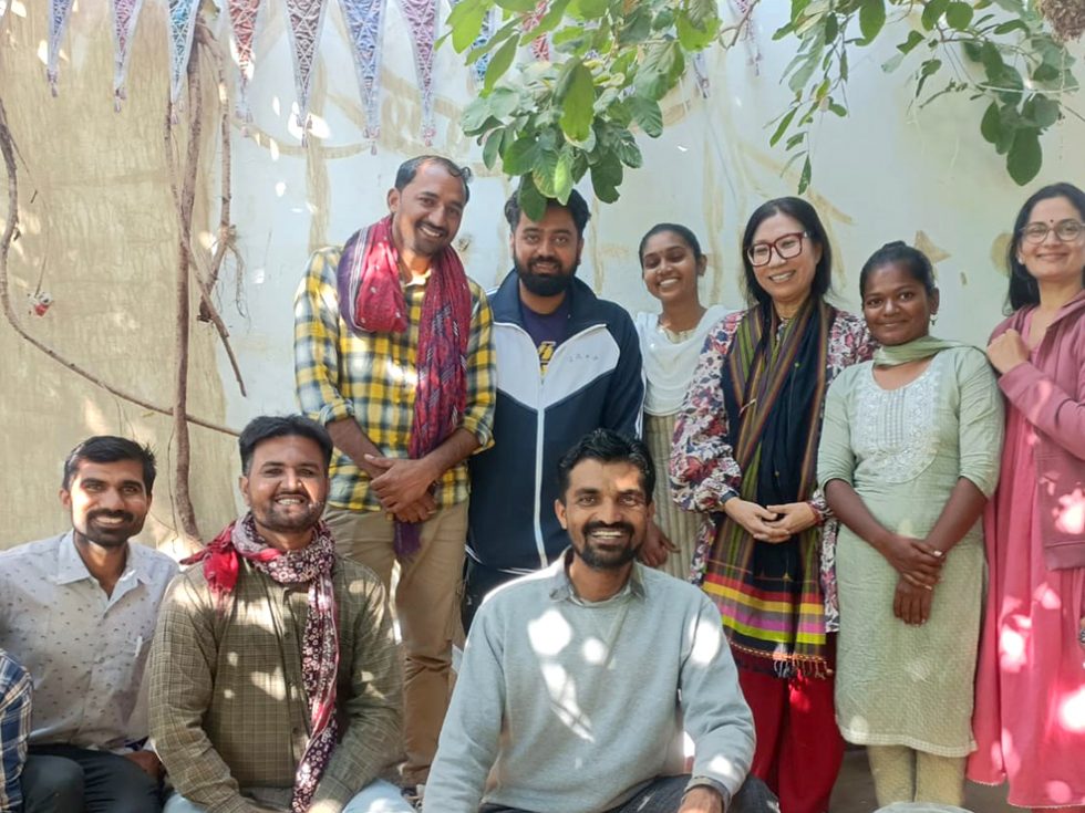 Professor Hanna Kim, PhD (fourth from the right, wearing eyeglasses and traditional Hindu garb), stands under a tree and in front of a mural of a tree, along with six men and four women, some of whom also are wearing Hindu apparel or scarves.