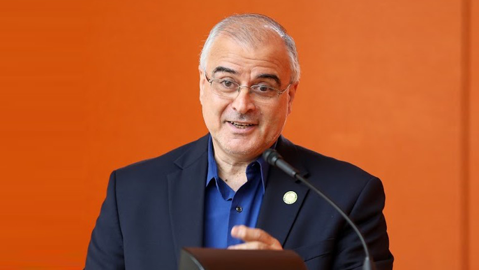 A man in a blue shirt and blazer stands in front of a microphone at a podium, smiling as he speaks to the audience.