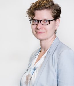 A woman with short brown hair and glasses smiling slightly. She wears a light-blue blazer and white blouse with floral print and is against a white background.