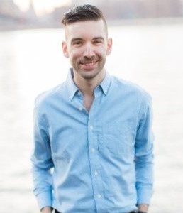 A man with brown hair grinning. He wears a blue button up shirt against a blurred background.