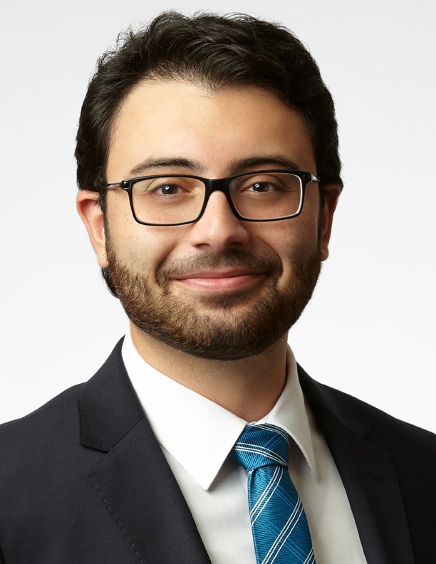 A headshot of a man with dark brown hair and glasses with a slight smile. He wears a white button up with a blue tie and a black blazer.