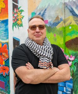  A man wearing sunglasses, black and white scarf, and black shirt, arms folded in front of a wall mural