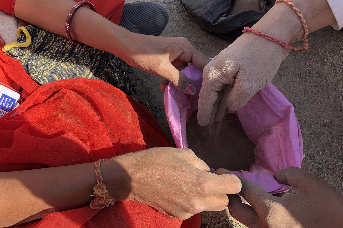 Three hands hold open a pink bag while another hand is pouring soil into the bag.