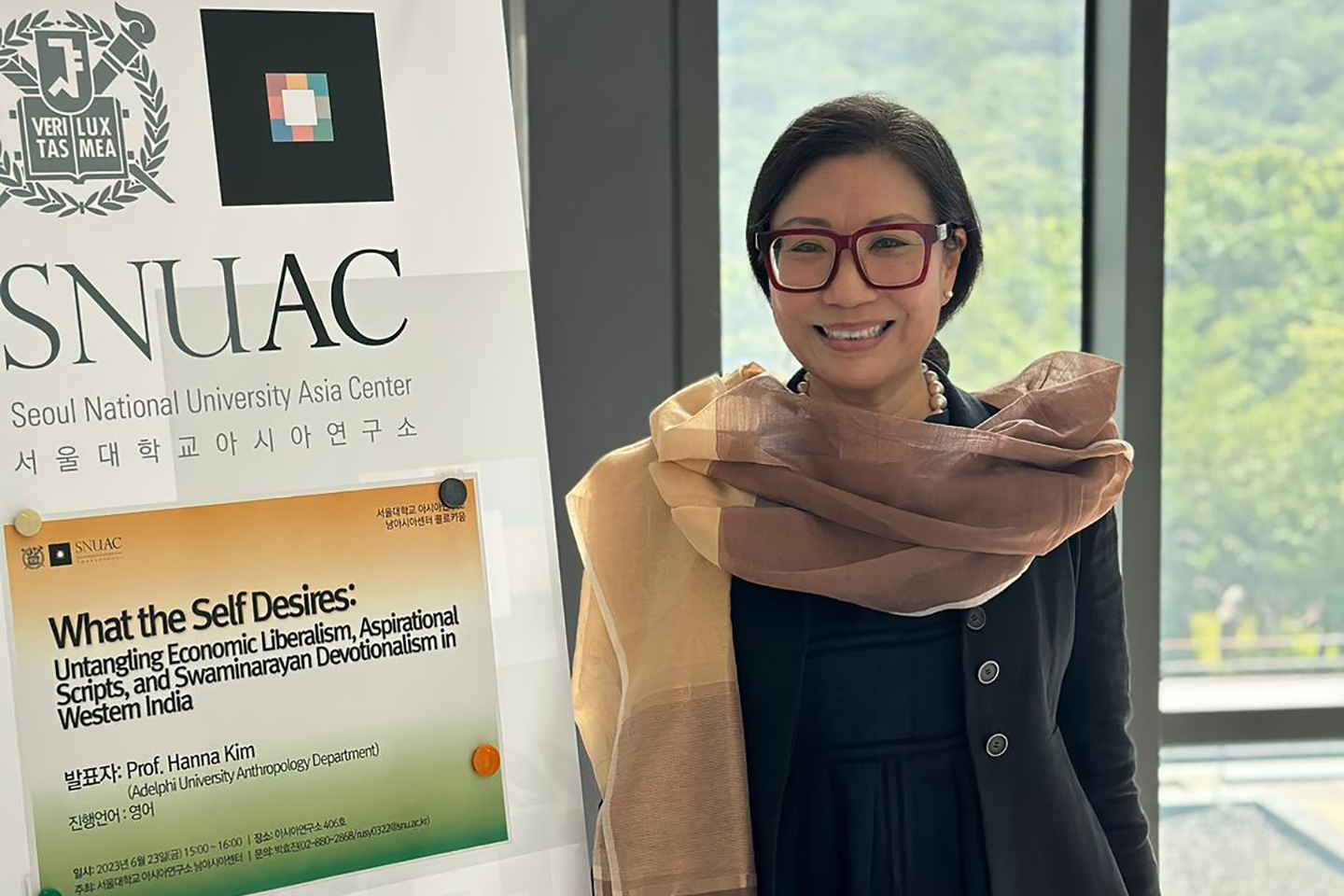 Professor Hanna Kim, wearing large red framed glasses stands to the right in front of a window. She is smiling. Next to her is a large sign containing a description of a led discussion.