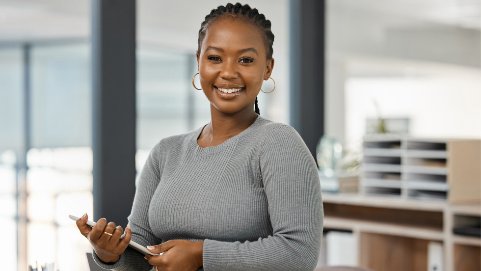 Standing in an office, a Black female in a gray sweater with short braids and hoop earrings holds a notepad.