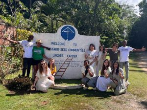 A group of 10 speech-language pathology students, during a Ruth S. Ammon College of Education and Health Sciences service learning trip to Jamaica, pose in front of a sign for Mustard Seed Communities, with trees in the background and a dirt road on the 