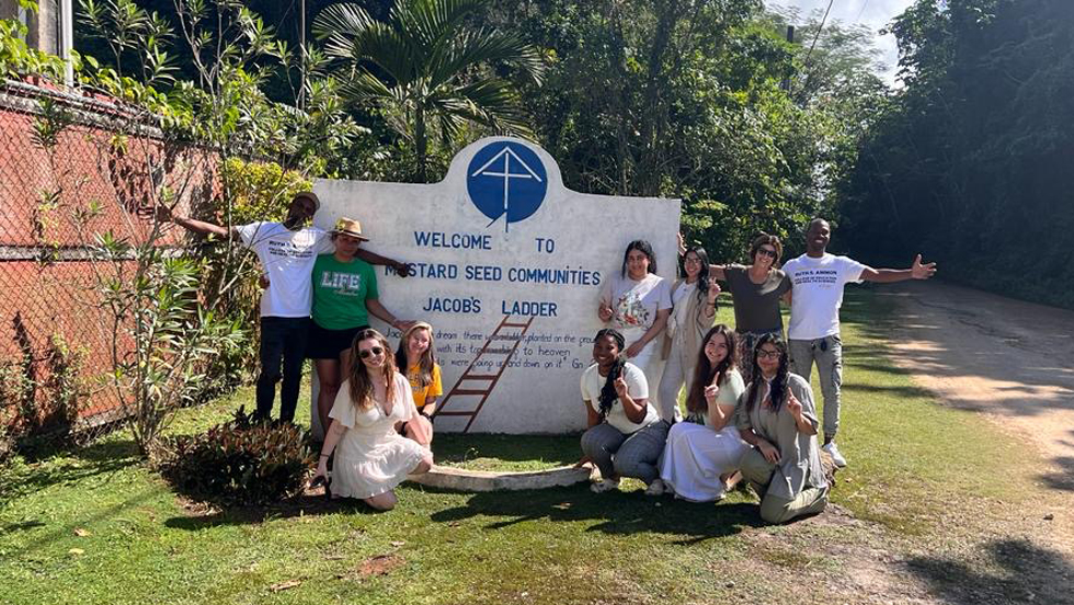 A group of 10 speech-language pathology students, during a Ruth S. Ammon College of Education and Health Sciences service learning trip to Jamaica, pose in front of a sign for Mustard Seed Communities, with trees in the background and a dirt road on the 