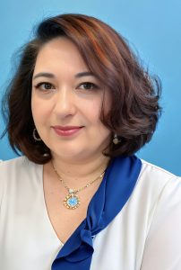 A woman, smiling slightly. She has a brown bob, earrings, a gold and sapphire necklace, white shirt, and blue scarf.