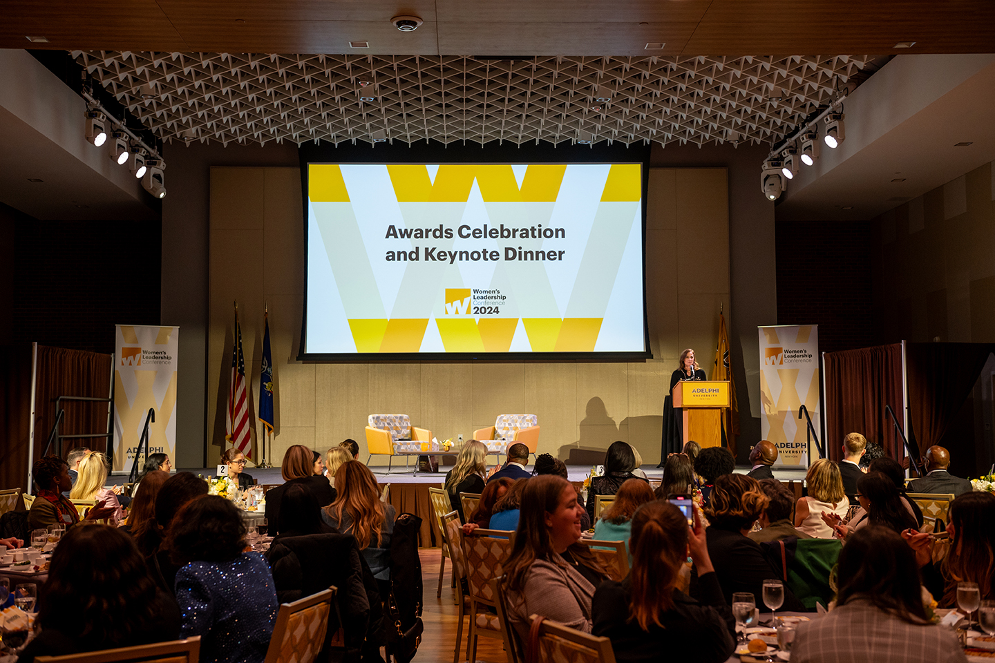 Woman stands at podium on the side of a stage. She is addressing a large crowd that's seated. Screen behind her reads "Awards Celebration and Keynote Dinner."