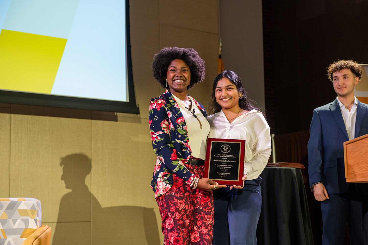 Two young women hold up plaque. Woman on the left is dressed in colorful florals. Woman on the right is dressed in a button-up shirt and slacks. A young man in a suit stands off to the right.