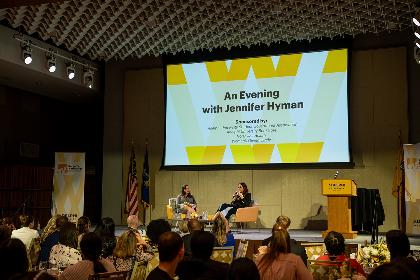 Two women sitting on stage appear to be talking to one another. A crowd of men and women are faced toward them. Screen behind two women reads "An Evening With Jennifer Hyman."