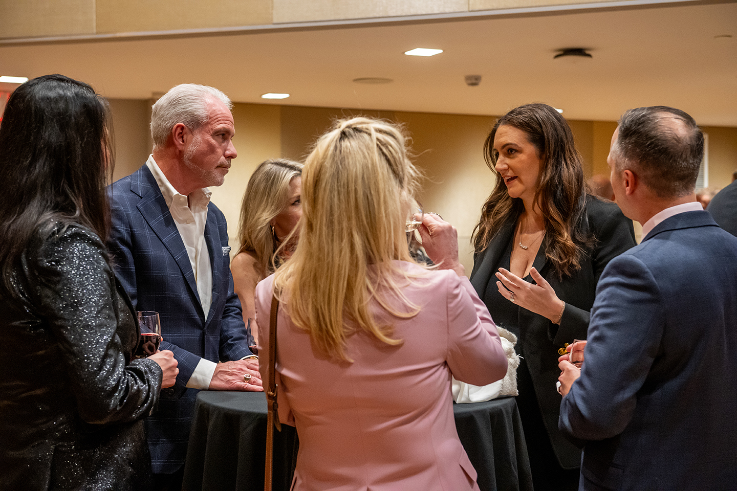 A group of women and men surrounds a high table. They hold glasses of wine. Woman with brown hair and black blazer appears to be actively speaking.