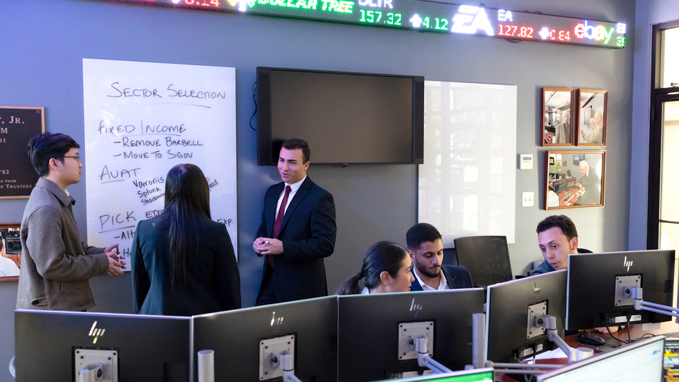 An instructor in a dark suit is talking to two college students. In front of them is a whiteboard with the words: "Sector Selection. Fixed Income" and other words that are illegible. Around them are students sitting in front of computer terminals.