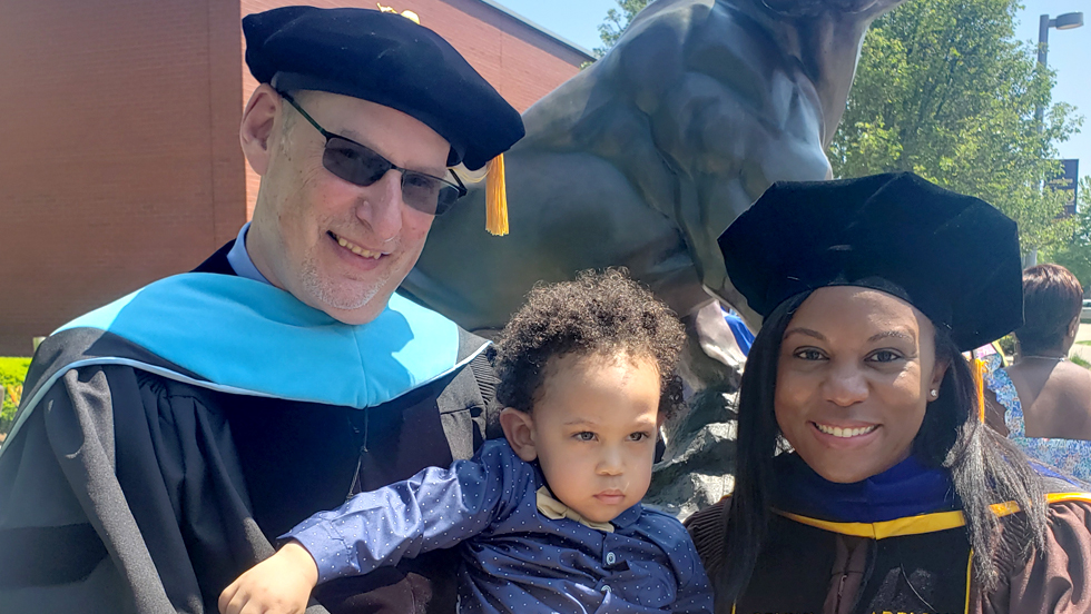  A white man wearing sunglasses and academic regalia stands next to a Black woman, also wearing academic regalia, with a toddler between them.