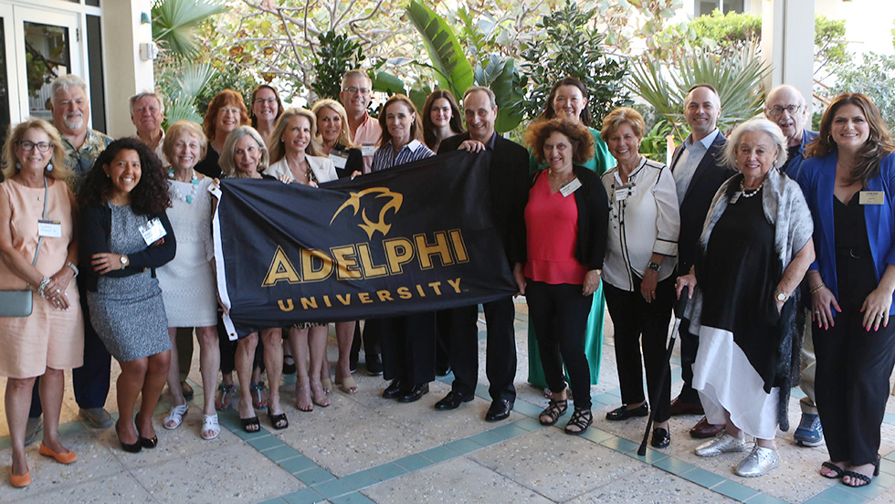 A group photo of attendees at a Momentum tour stop, taken on the patio surrounded by tropical flora.  There are more than 20 people in the photo, including President Riordan.  