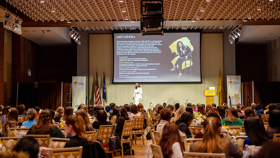 Kendra Bracken-Ferguson addresses attendees from the stage. Behind her is a digital screen that says "Meet Kendra" and includes her photo and a short biography.