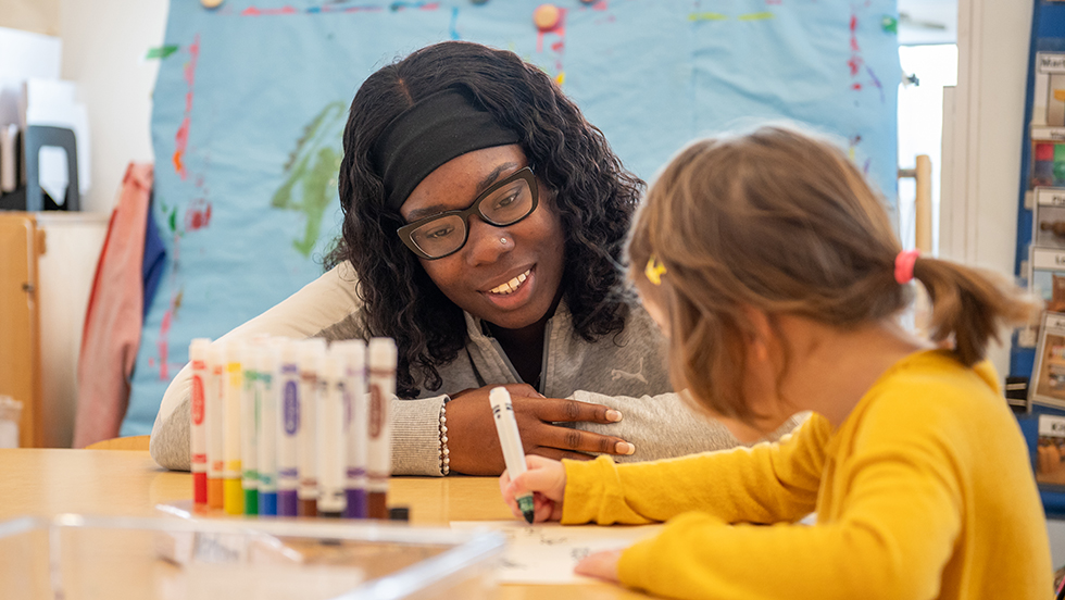  An adult with glasses and a headband observes a young child drawing with markers at a table in a classroom setting.