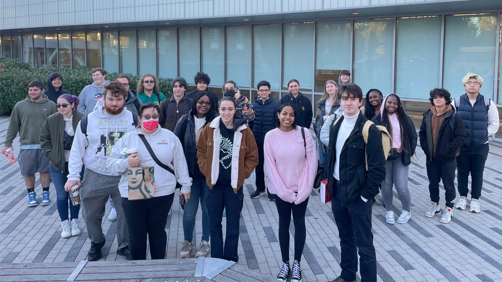 About 20 college students, of various ethnicities, wearing casual clothing, in front of a building