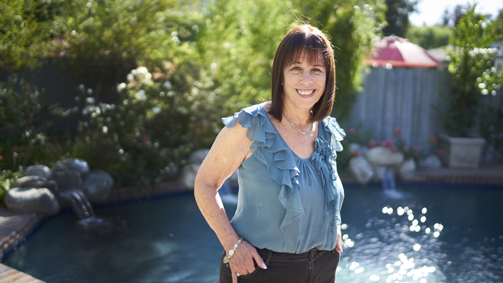 A smiling woman with long brown hair wearing a frilly sleeveless blue blouse stands before a pool with blue water, with green bushes in the background.