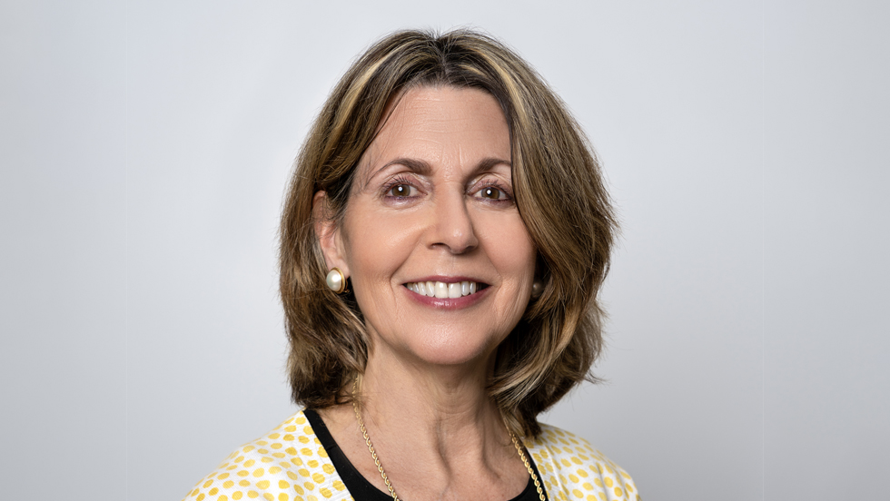 A smiling woman, whose long brown hair has blonde highlights, is wearing pearl earrings, a gold necklace and a yellow polka dot jacket.