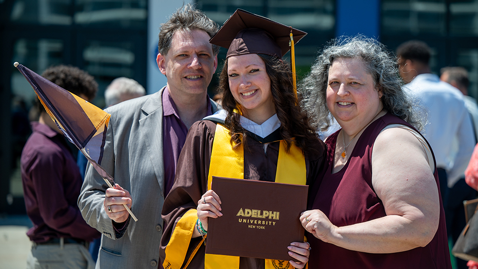 Young woman in cap and gown holds diploma between an older woman and man.