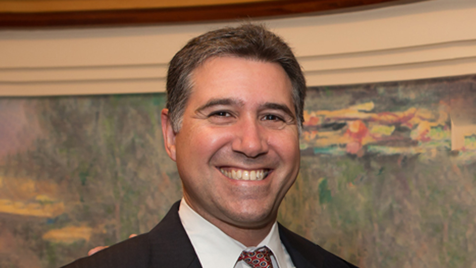 A smiling man with short brown hair and graying sideburns, wearing a dark suit, white shirt and polka dot tie, stands before a painting. 