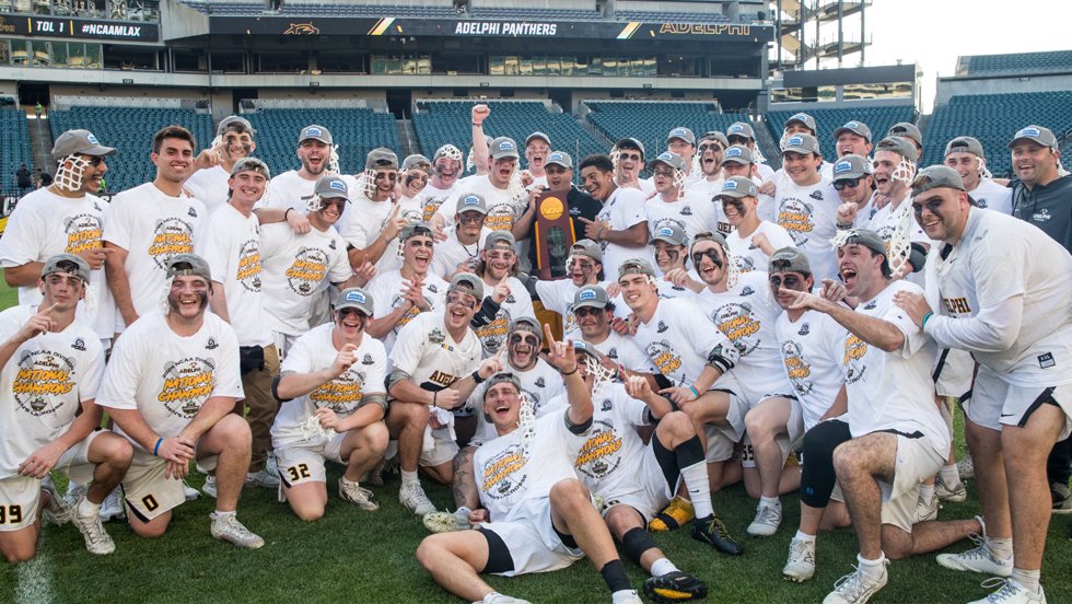 A large group of athletes in white shirts and caps gather on a field, celebrating with a trophy in the center. 