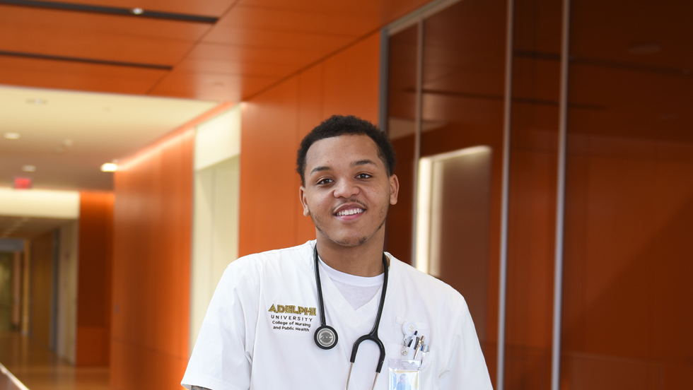 A Black male nursing student, wearing white scrubs and with a stethoscope draped around his neck, smiles as he stands in a corridor. 
