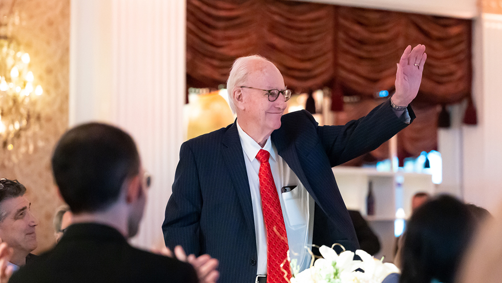 An older, white-haired man with eyeglasses and wearing a pinstriped suit and. red necktie stands and waves with his left arm, as men seated nearby applaud.