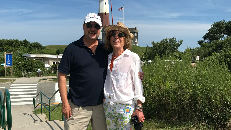 A man and woman are standing before a lighthouse and greenery. She is wearing sunglasses, a straw hat, a long-sleeved white blouse and print slacks, while he is wearing sunglasses, a white cap, a short-sleeved black shirt and beige slacks. 