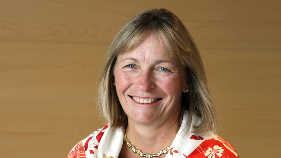 A smiling woman with long light brown hair and blonde highlights, is wearing earrings, a gold necklace and a red-and-white print blouse. 