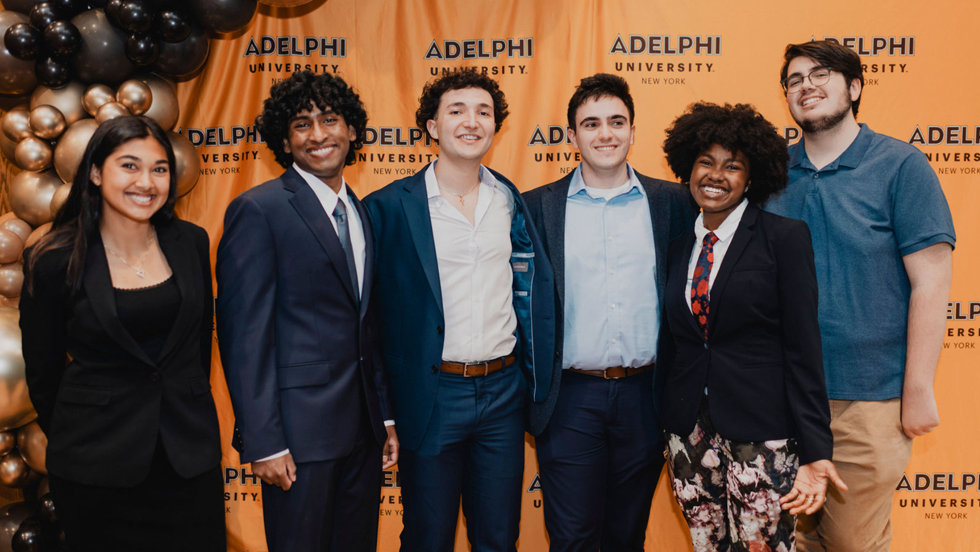  Six students of different genders and ethnicities smile facing the camera. There are brown and gold balloons on the left and they stand in front of a gold background with the words “Adelphi University.”