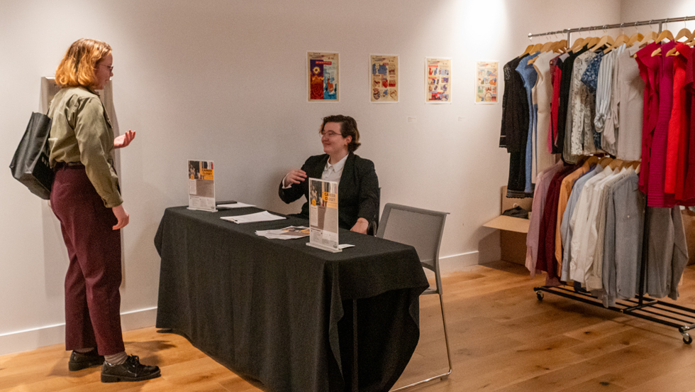 Woman greeting someone in a suit jacket who's sitting at a table with a clothes rack behind them