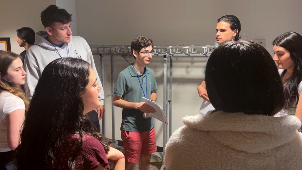 A college student in a room is speaking toward his classmates standing around him.