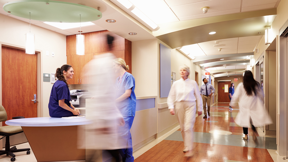 Two female nurses in blue scrubs chat by the nurses station while five hospital staffers walk up and down the brightly lit corridor. 