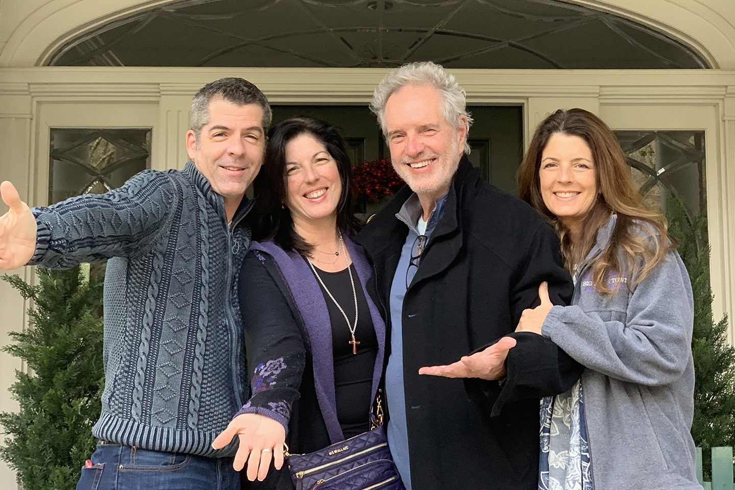 Two men and two women stand together, the first three from left each with one arm out, all smiling