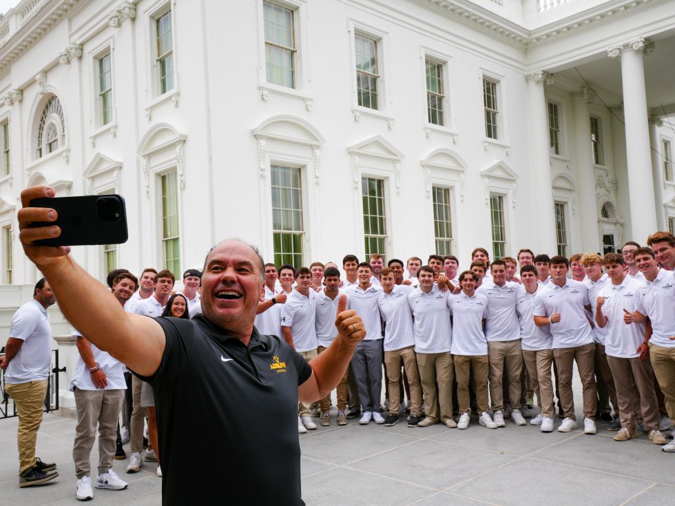 The Adelphi men's lacrosse coach holds his phone out in front of him to take a selfie with Adelphi student-athletes in front of the White House in Washington, D.C. 