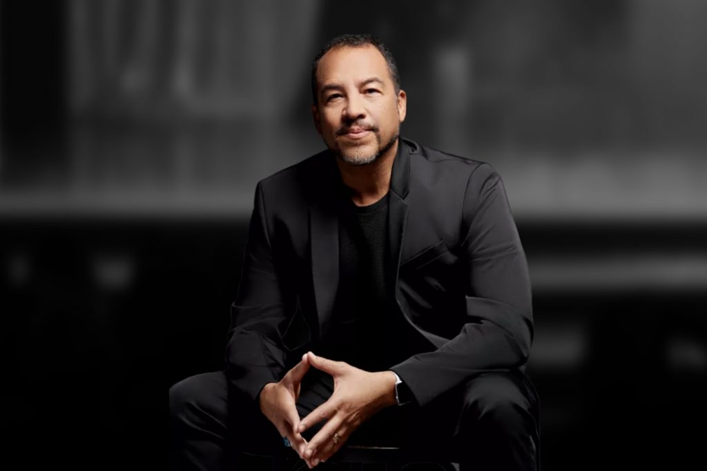 A Hispanic man dressed in black. He is seated and smiling slightly.