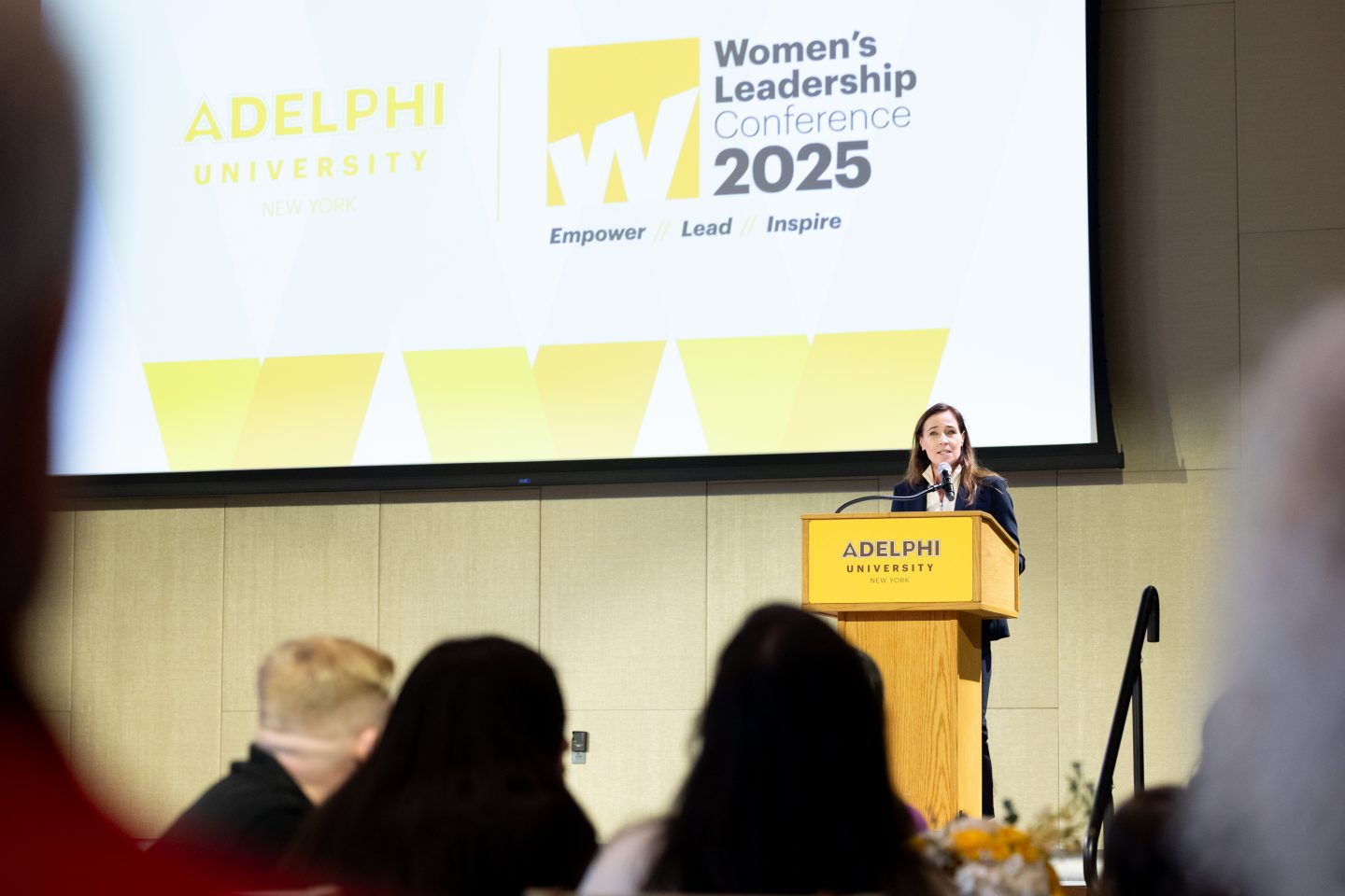 President Christine Riordan at a lectern in front of a large sign that says "Women's Leadership Conference 2025" (a dais would be seating at a table in the front)