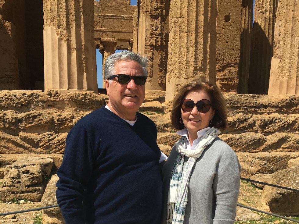 Man in blue sweater stands next to his wife in a gray sweater - both wearing sunglasses -  in front of Greek ruins.