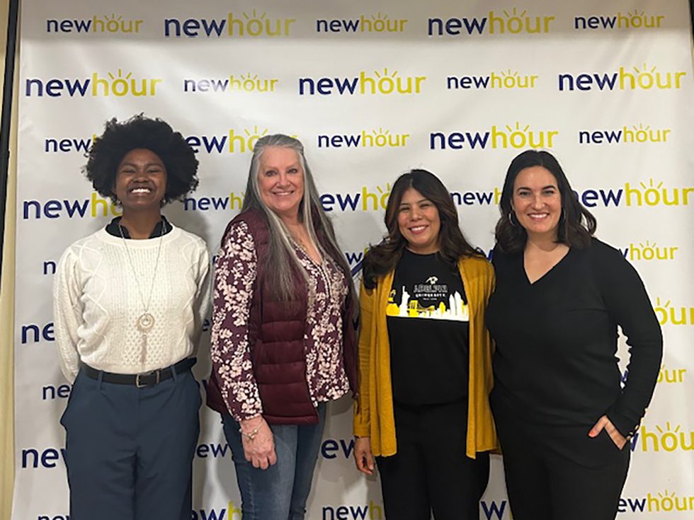 Four women posing and smiling in front of a step and repeat background that says New Hour