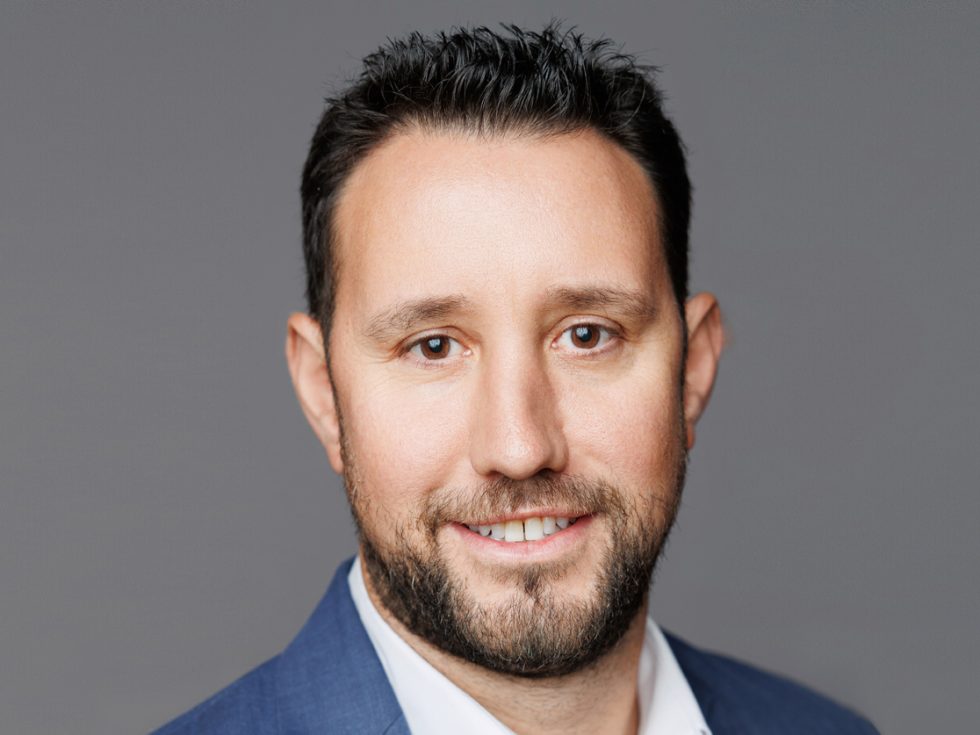 Headshot of a man with short dark hair and a trimmed beard, wearing a blue suit jacket and white collared shirt, smiling slightly against a plain gray background